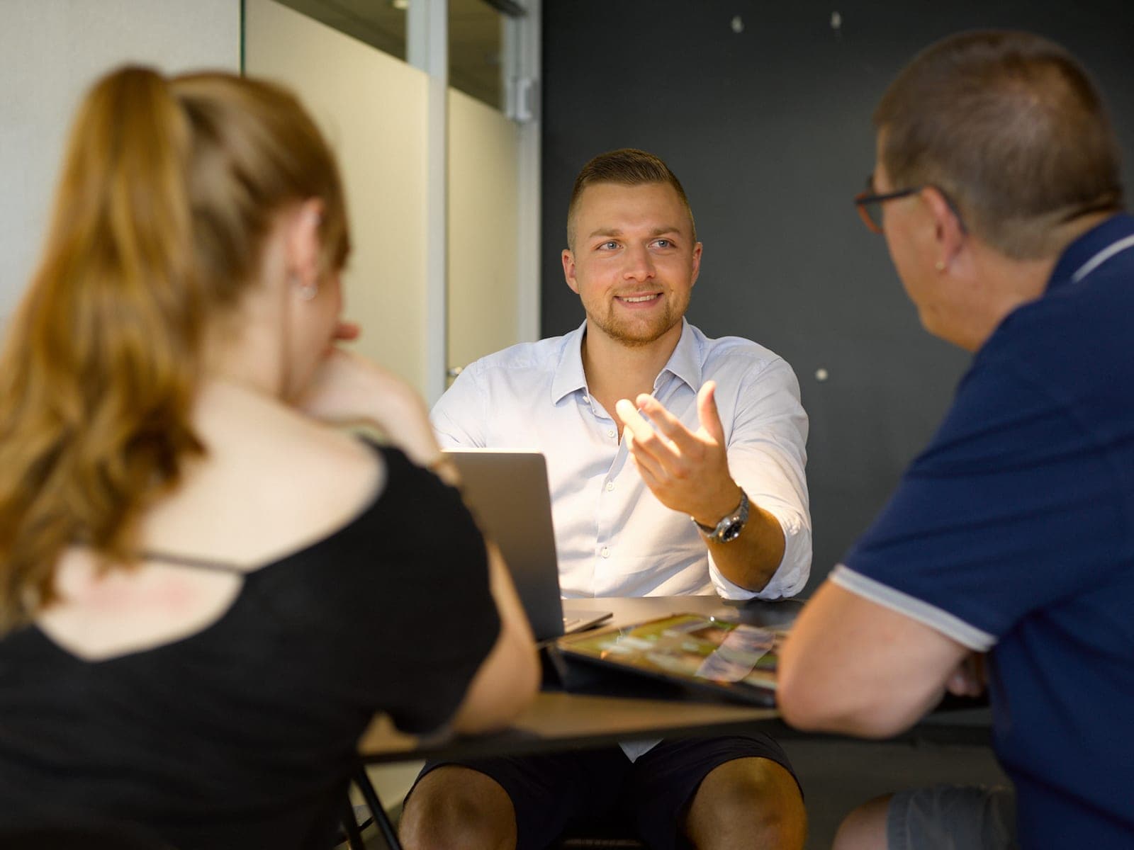 Besprechung an einem Tisch: ein Mitarbeiter erklärt etwas, vor ihm ein aufgeklapptes Notebook